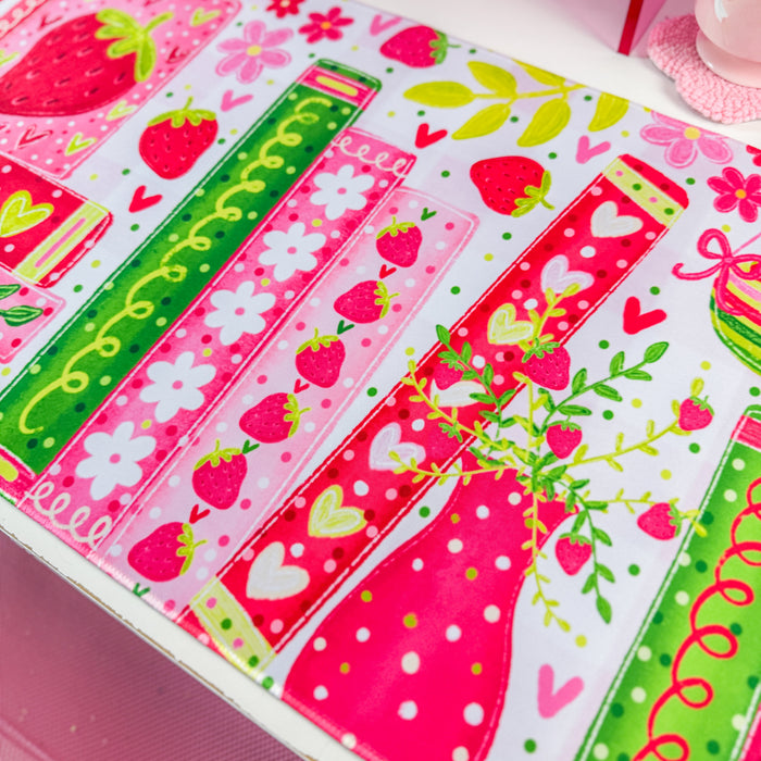A close-up of a colorful printed fabric or mat featuring strawberry motifs, pink and green stripes, hearts, flowers, and dotted patterns on a white base.