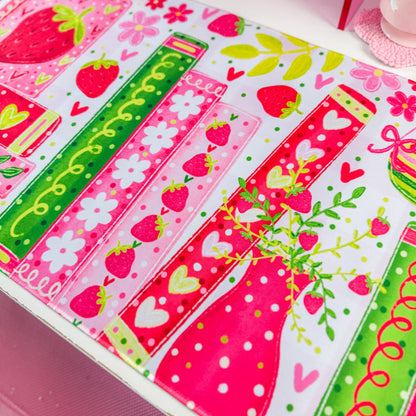A close-up of a colorful printed fabric or mat featuring strawberry motifs, pink and green stripes, hearts, flowers, and dotted patterns on a white base.