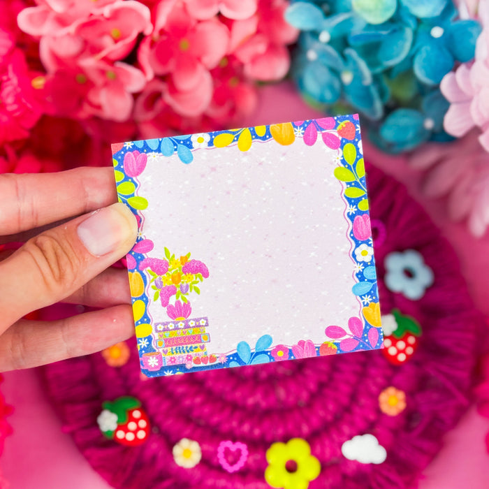 Hand holding sticky notes featuring a floral design with a book stack, and a border of vines and florals, on a pink backdrop.