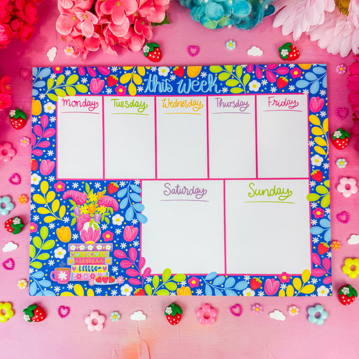 A weekly planner pad with a colorful floral border and white writing sections for Monday through Sunday, styled on a pink background with flowers and small decorative accents.