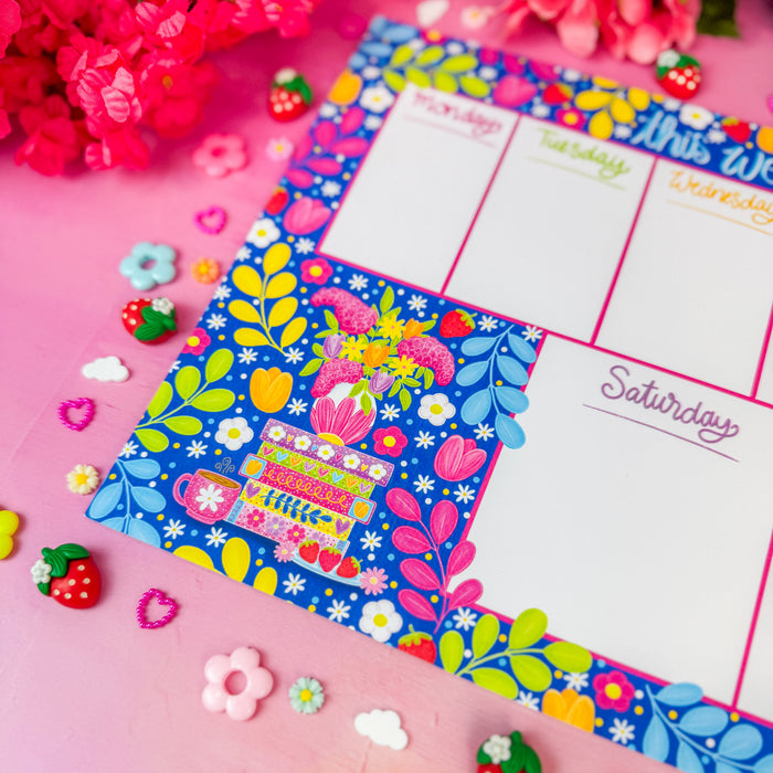 A colorful weekly planner page with floral borders and labeled days of the week, styled on a pink surface with flowers, small flower charms, and strawberry accents.