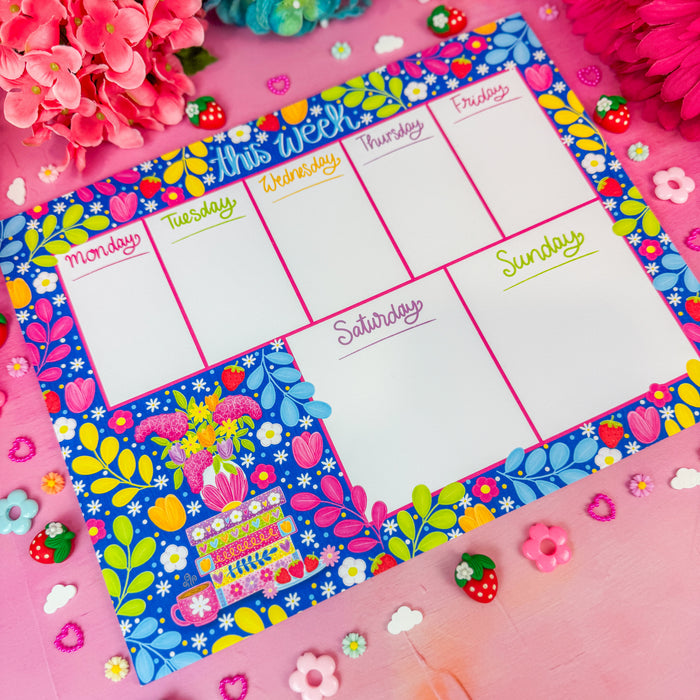 A weekly planner pad with a colorful floral border and white writing sections for Monday through Sunday, styled on a pink surface with flowers and small decorative accessories around it.