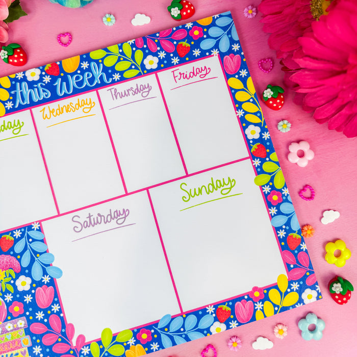 A weekly planner notepad with a colorful floral border, laid flat on a pink background surrounded by flowers and small decorative strawberries.