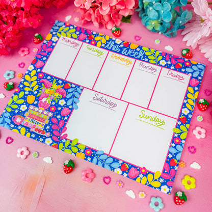 A colorful weekly planner pad with a vibrant floral border and white writing sections, styled on a pink surface with decorative flowers and small strawberry embellishments.