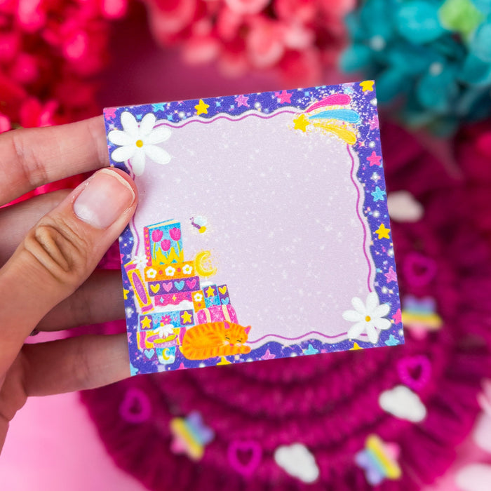 A hand holds a small square notepad with a colorful border featuring a rainbow, stars, flowers, and a decorated cake illustration. The notepad has a light pink center with subtle glitter speckles.