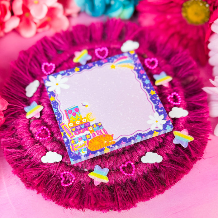 A circular hot pink faux fur coaster with a fringe edge, decorated with small colorful stickers including hearts, clouds, and a cartoon cat on a blue border. A sticky notepad is placed in the center.