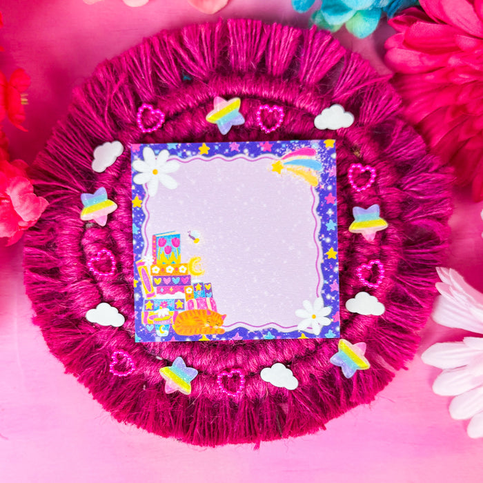 A round magenta fringe decorative coaster with a  sticky notepad featuring a colorful cartoon book stack with a cat illustration in the lower-left corner, surrounded by small white clouds, hearts, stars, and butterflies on a pink background with flowers around the edges.